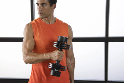 A man in an orange sleeveless athletic shirt holding a black adjustable dumbbell in front of a white window with black frames.
