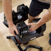 A close-up of a person adjusting black adjustable dumbbells on a weight bench in a home gym setting with a light wood floor.