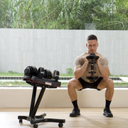 A man in a home gym setting performing a squat with a dumbbell, with a weight bench, a dumbbell rack, and wooden blocks in the background.