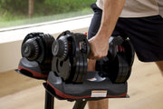 A person in a gym setting holding a 66lb adjustable dumbbell from a stack of weights on a stand. The dumbbells are black with red accents and white printed markings.