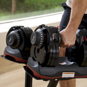 A person in a gym setting holding a 66lb adjustable dumbbell from a stack of weights on a stand. The dumbbells are black with red accents and white printed markings.