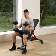 A man sits on a black and red adjustable weight bench in a bright gym, holding dumbbells with red accents.