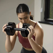 A woman in a maroon sports bra and dark gray leggings holds a black adjustable dumbbell with silver plates in front of her chest in an indoor setting.