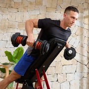 A man in a black t-shirt and blue shorts performs a shoulder press on a black adjustable weight bench with red legs, using dumbbells against a stone wall background.