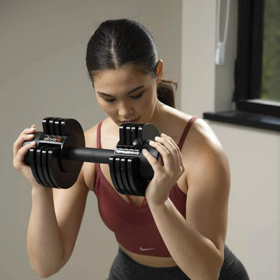 A woman in a maroon sports bra and dark gray leggings holds a black adjustable dumbbell with silver plates in front of her chest in an indoor setting.