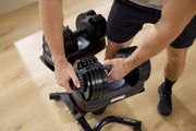 A close-up of a person adjusting black adjustable dumbbells on a weight bench in a home gym setting with a light wood floor.