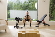 A man in a home gym setting performing a squat with a dumbbell, with a weight bench, a dumbbell rack, and wooden blocks in the background.