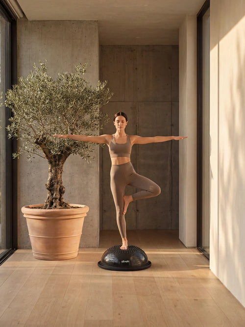 Woman practicing yoga on a balance ball in a modern indoor setting with a potted olive tree.