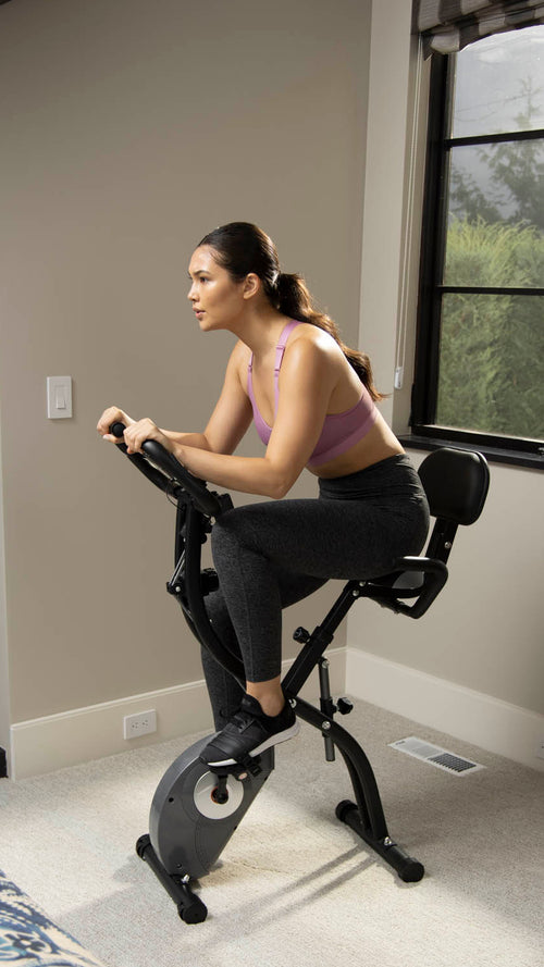 A woman exercising on a black indoor stationary bike in a bedroom with beige walls, windows, and a patterned bedspread visible in the foreground.