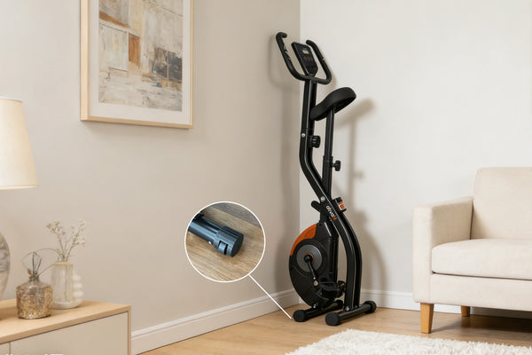 A compact upright exercise bike with a black frame and seat, orange side accents, and a large dark flywheel, shown in a bright living room corner next to a sofa. A circular inset highlights the bike's compact storage footprint on a wooden floor.