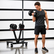 A man in a gym setting holds a black adjustable dumbbell while standing next to a black weight bench with two more dumbbells on a wheeled cart.