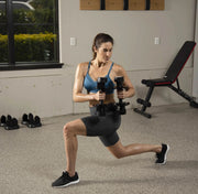 A woman in a blue sports bra and black shorts performs a lunge while holding dumbbells in a home gym setting with a weight bench and exercise shoes visible.