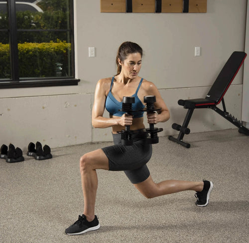 A woman in a blue sports bra and black shorts performs a lunge while holding dumbbells in a home gym setting with a weight bench and exercise shoes visible.
