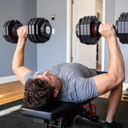 A man lying on a weight bench doing a chest press with black adjustable dumbbells in a home gym setting.