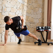A man in a black t-shirt and blue shorts performs a dumbbell side plank exercise in a gym with a stone wall background, with a dumbbell rack visible behind him.